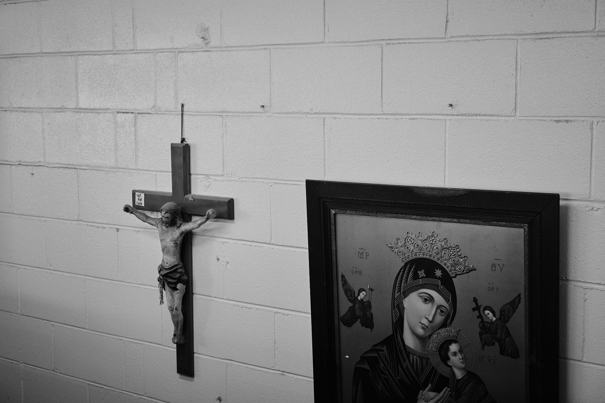 A statue of a crucified Jesus hangs on the wall beside a painting of the Virgin Mary. Upon the cross is a sticker that reads 'Not for sale'.